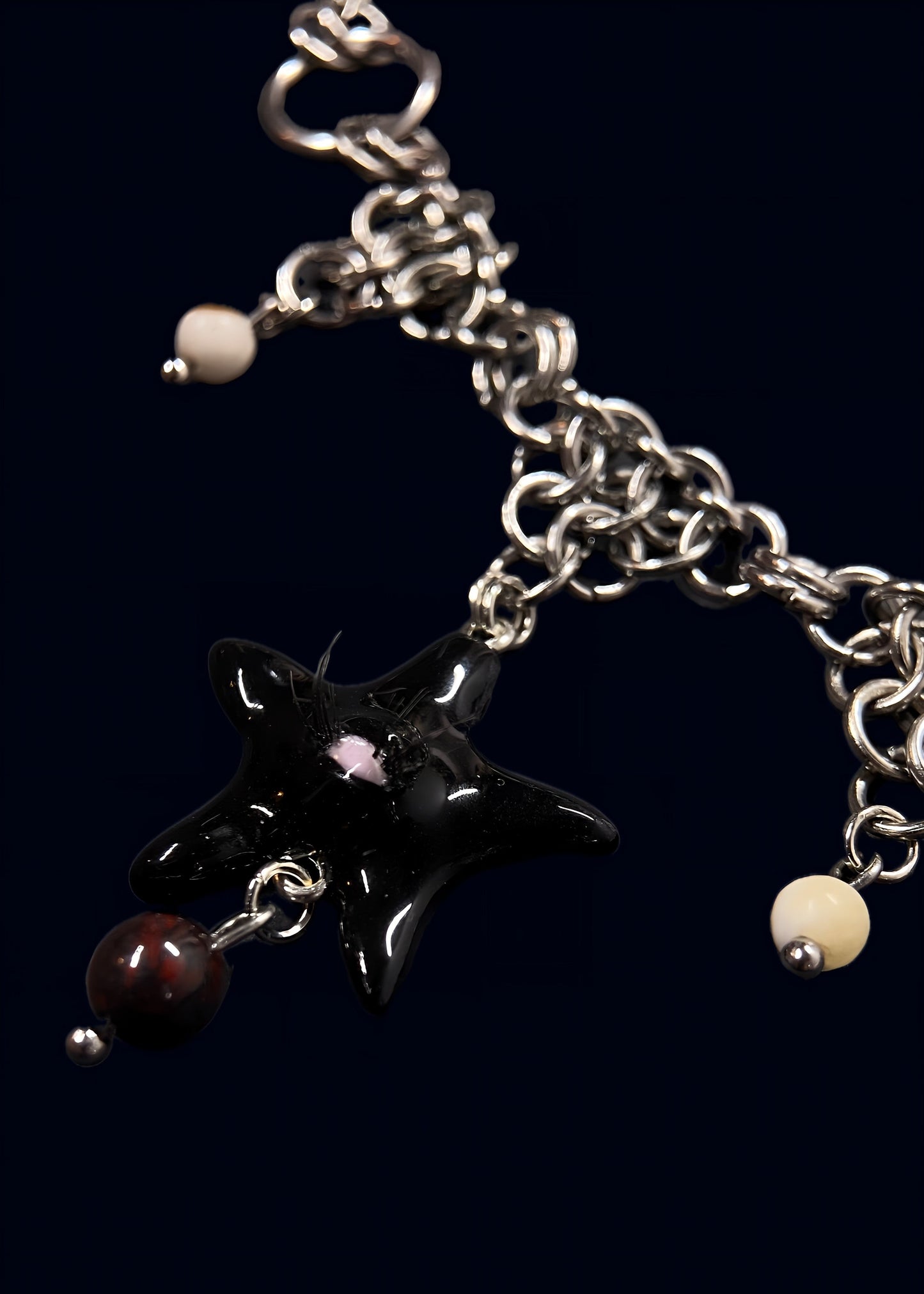 front close-up of black star pendant with pink eye detail and fine lashes, glossy resin surface, suspended from chunky silver chainmail with small bead charms