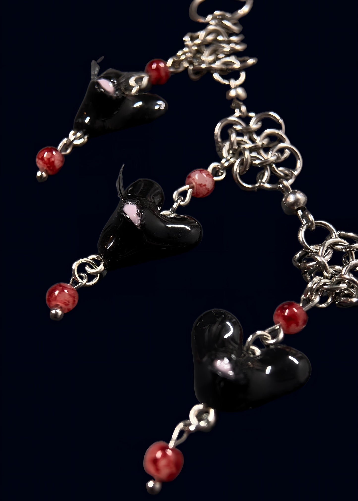 close up product photo of the other side of smaller pendants from handmade black heart swarm necklace. focus on small black heart pendant with one pink eye and small lashes, thrifted red pearls dangling from the heart and chainmail necklace. 2 more small pendants can be seen out of focus.