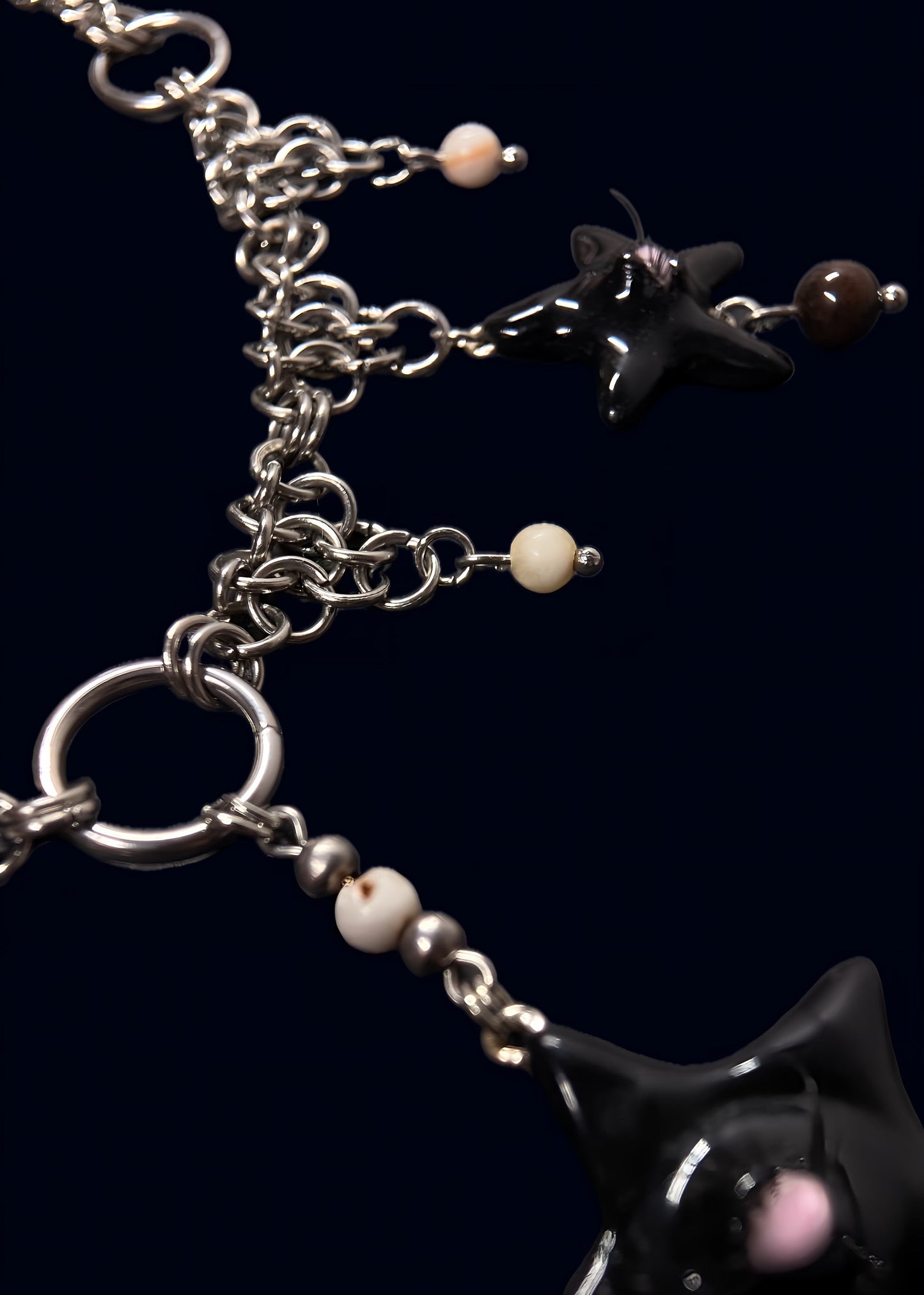 angled detail photo of black gazing star necklace showing metal rings, layered chainmail structure and small cream bead accents on dark blue background