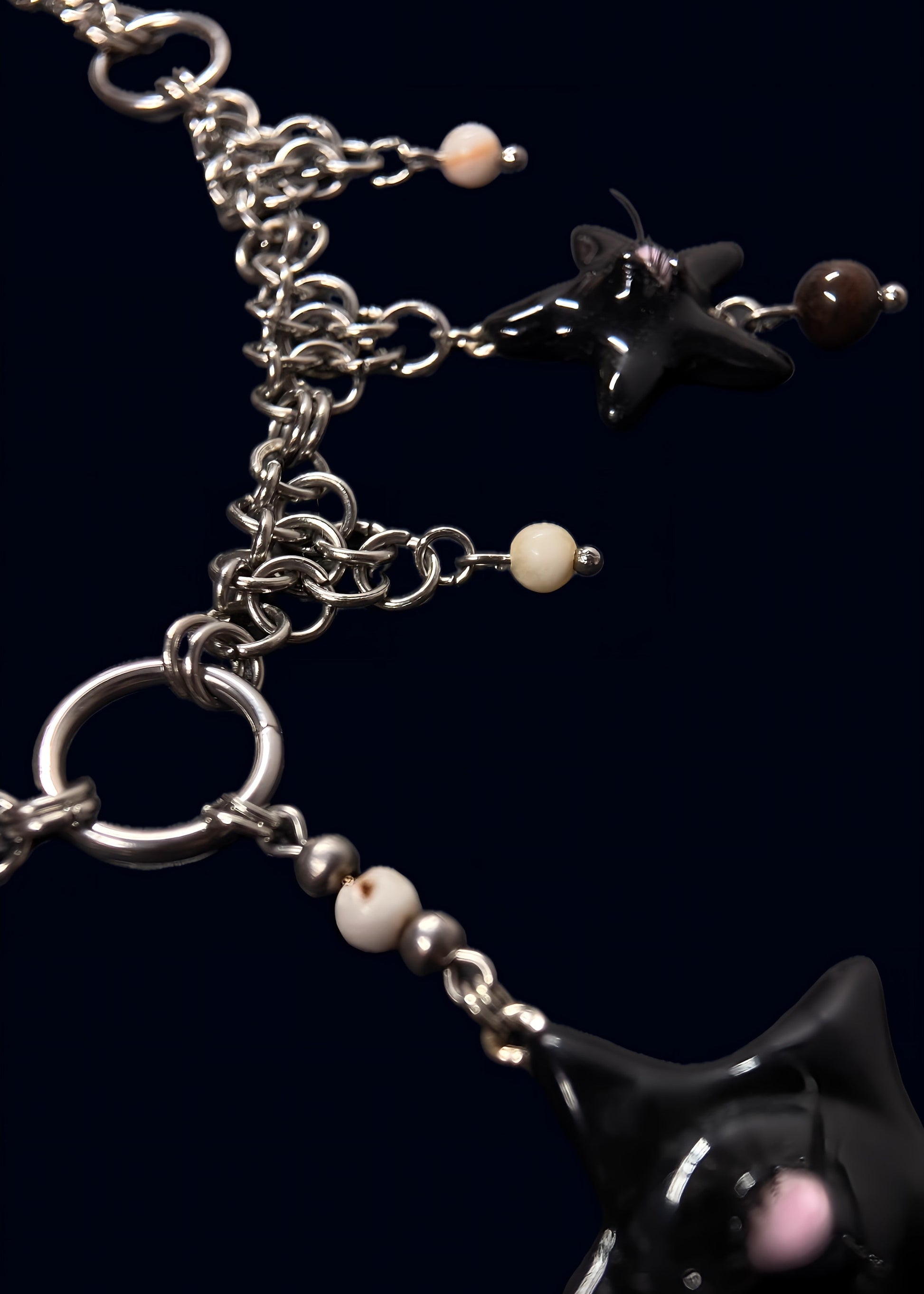 angled detail photo of black gazing star necklace showing metal rings, layered chainmail structure and small cream bead accents on dark blue background