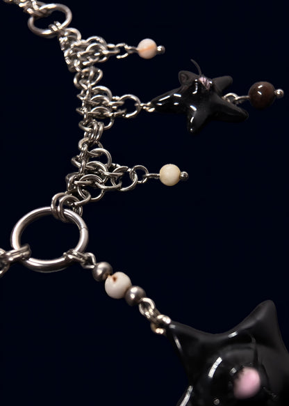angled detail photo of black gazing star necklace showing metal rings, layered chainmail structure and small cream bead accents on dark blue background