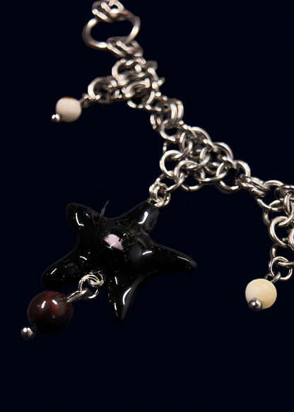 front close-up of black star pendant with pink eye detail and fine lashes, glossy resin surface, suspended from chunky silver chainmail with small bead charms