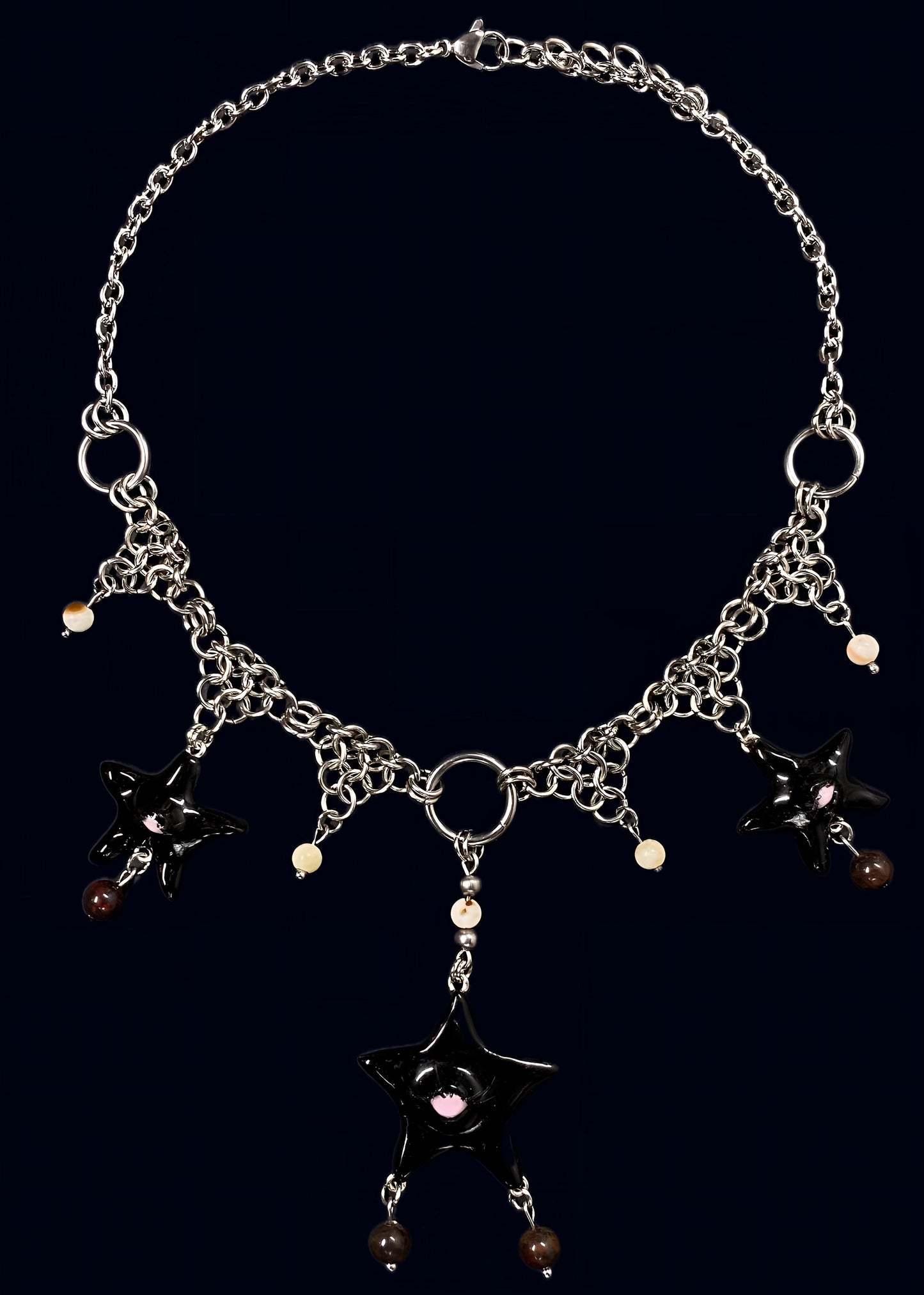full necklace view of black gazing star necklace laid flat on dark blue background, central black eye star pendant with dangling bead drops and symmetrical chainmail arrangement