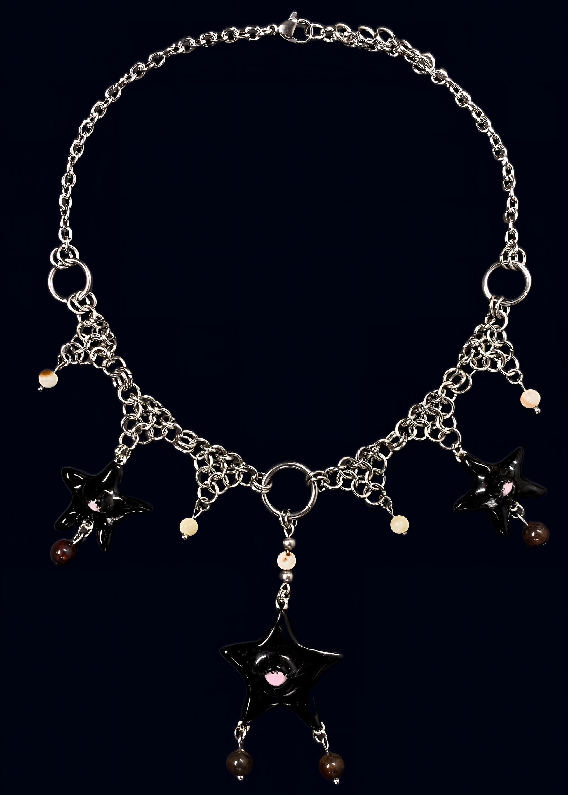 full necklace view of black gazing star necklace laid flat on dark blue background, central black eye star pendant with dangling bead drops and symmetrical chainmail arrangement