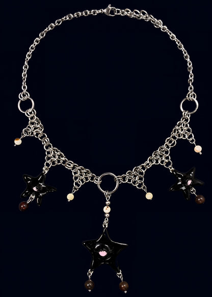 full necklace view of black gazing star necklace laid flat on dark blue background, central black eye star pendant with dangling bead drops and symmetrical chainmail arrangement