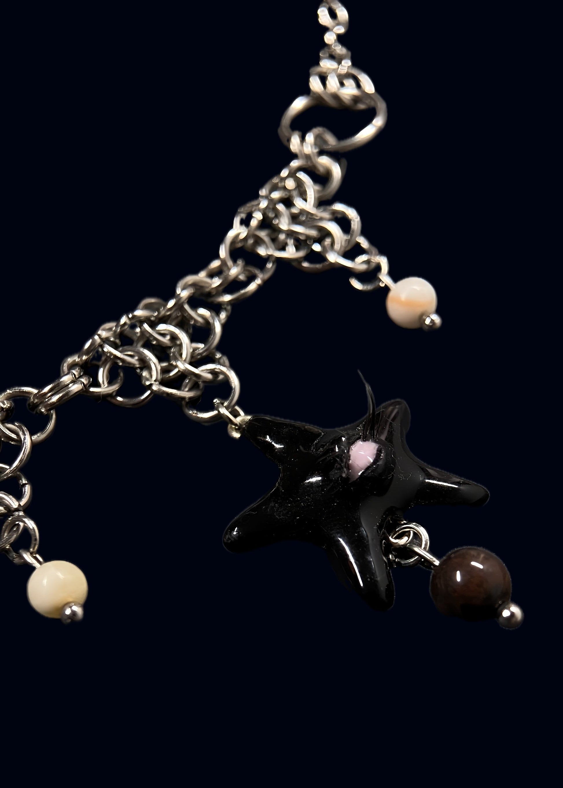 close-up product photo of black gazing star necklace on dark blue background. glossy black star pendant with single eye and lashes, attached to layered stainless steel chainmail with small cream and dark brown bead drops
