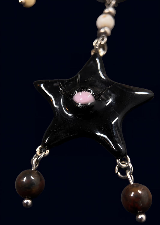 close-up of glossy black gazing star pendant with single eye and lashes, two dangling brown peals below, attached to silver chainmail on dark blue background