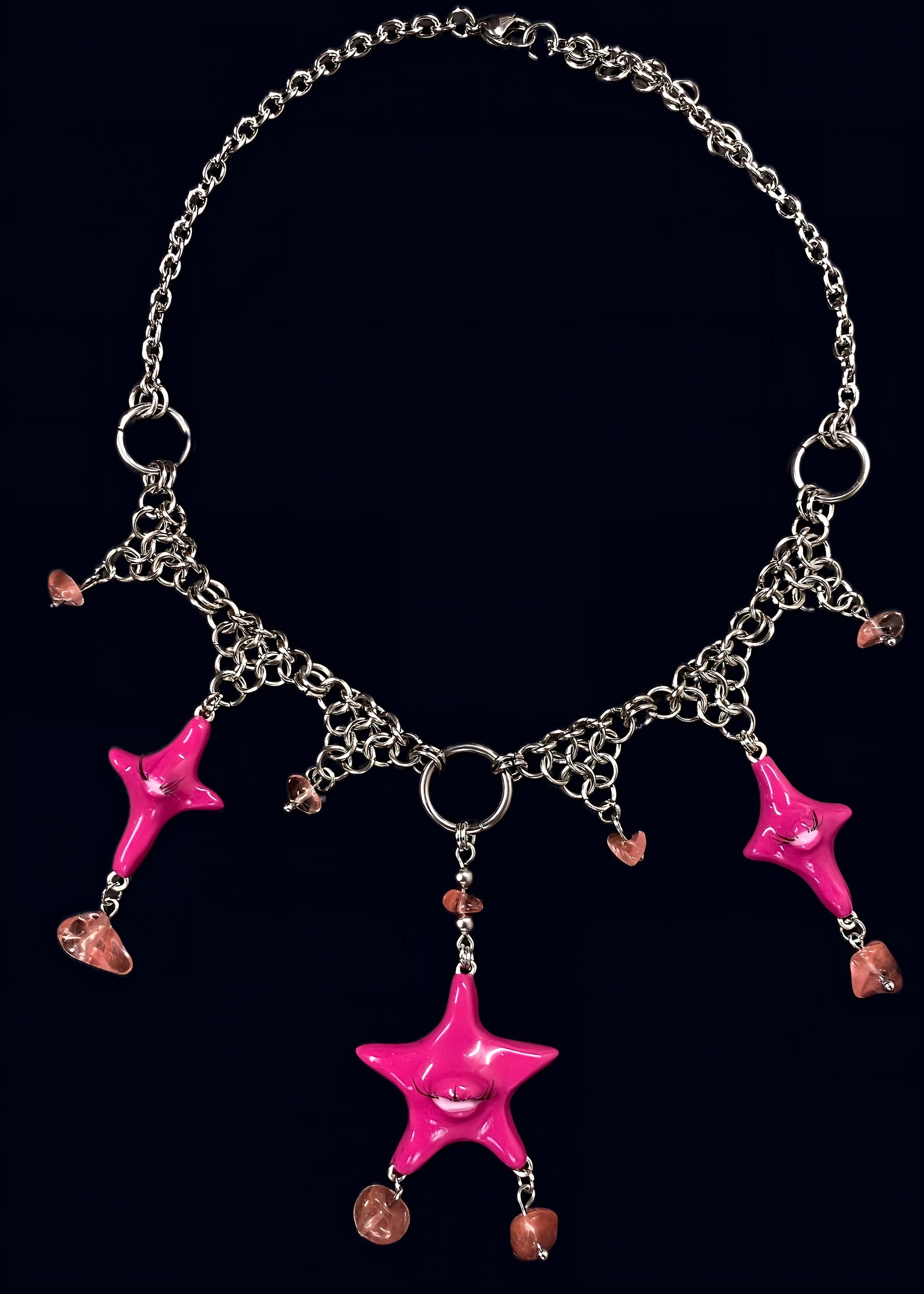 full necklace view of pink gazing star necklace laid flat on dark blue background, central pink eye star pendant with dangling gems drops and symmetrical chainmail arrangement