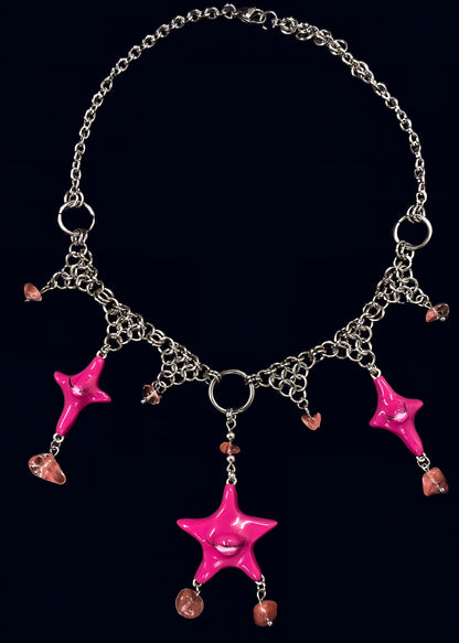 full necklace view of pink gazing star necklace laid flat on dark blue background, central pink eye star pendant with dangling gems drops and symmetrical chainmail arrangement