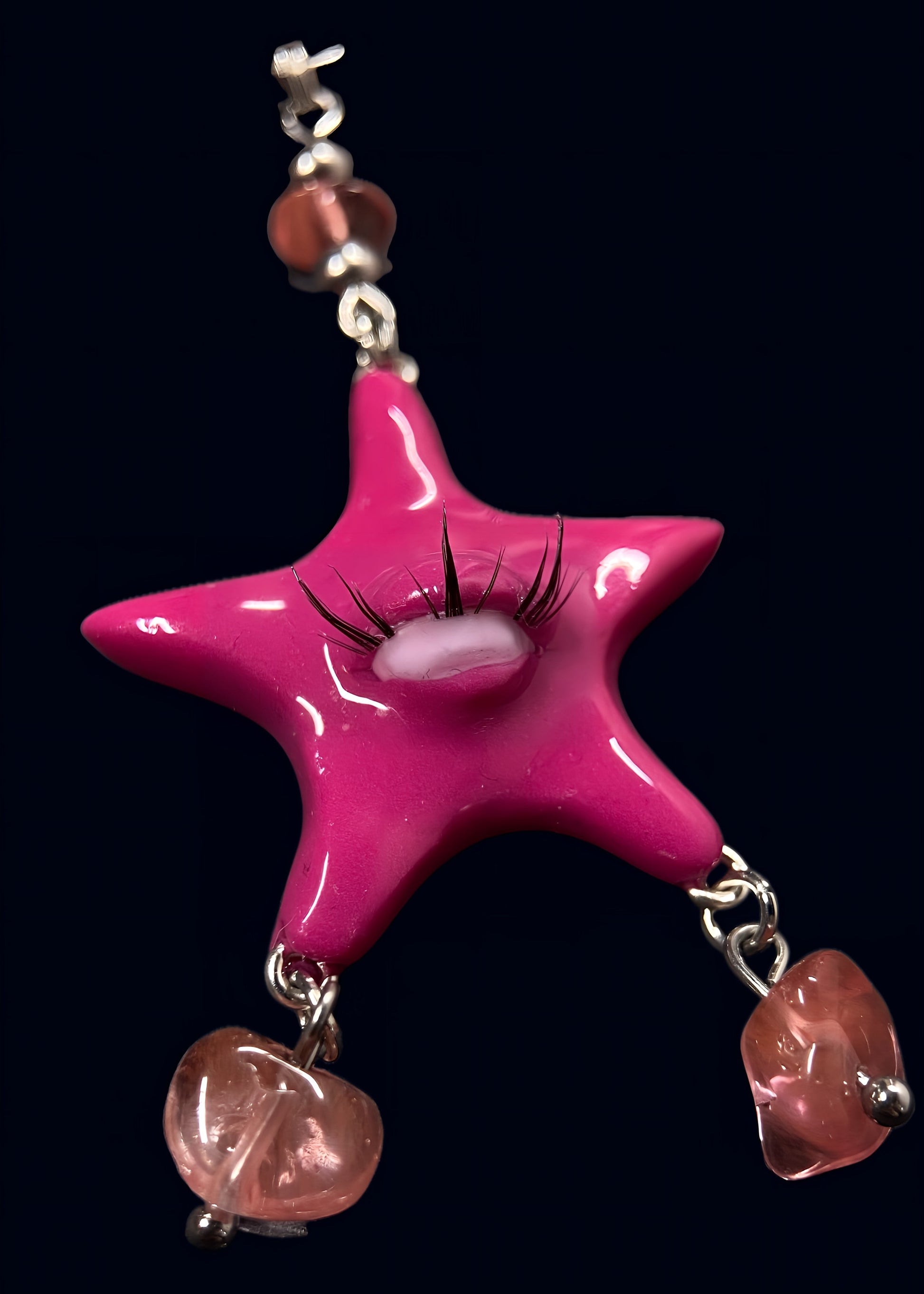 close-up of glossy pink gazing star pendant with single eye and lashes, two dangling rose quartz chip drops below, attached to silver chainmail on dark blue background