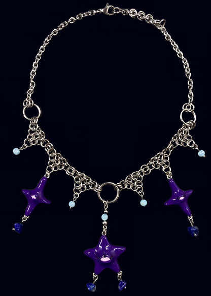 full view of purple gazing star necklace laid flat on dark blue background, central purple eye star pendant with multiple dangling stone drops and symmetrical chain arrangement