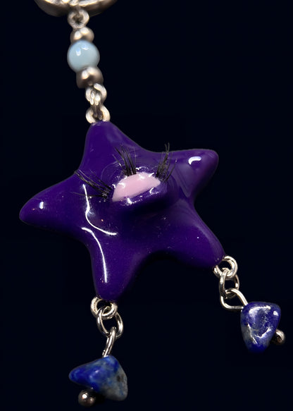 close-up of purple gazing star pendant with single eye and lashes, glossy resin finish, dangling dark blue stone chips below, attached to silver chainmail