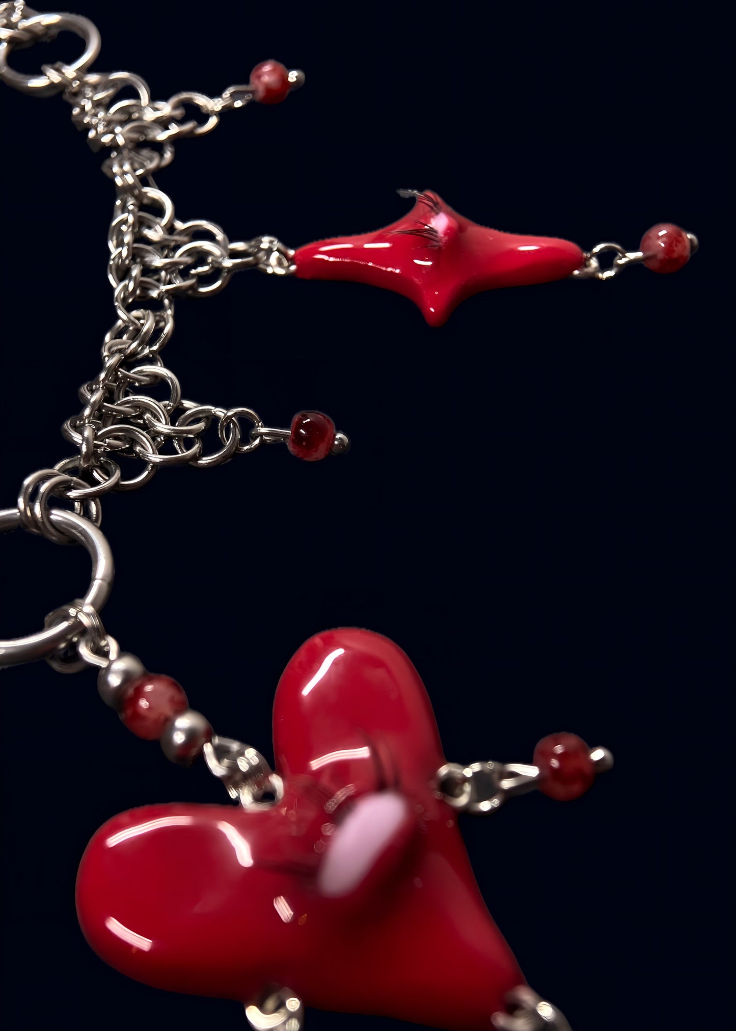 close-up of red heart creature necklace on dark blue background, glossy red heart pendant with single eye and lashes, red bead drops and small red star charm attached to silver chainmail