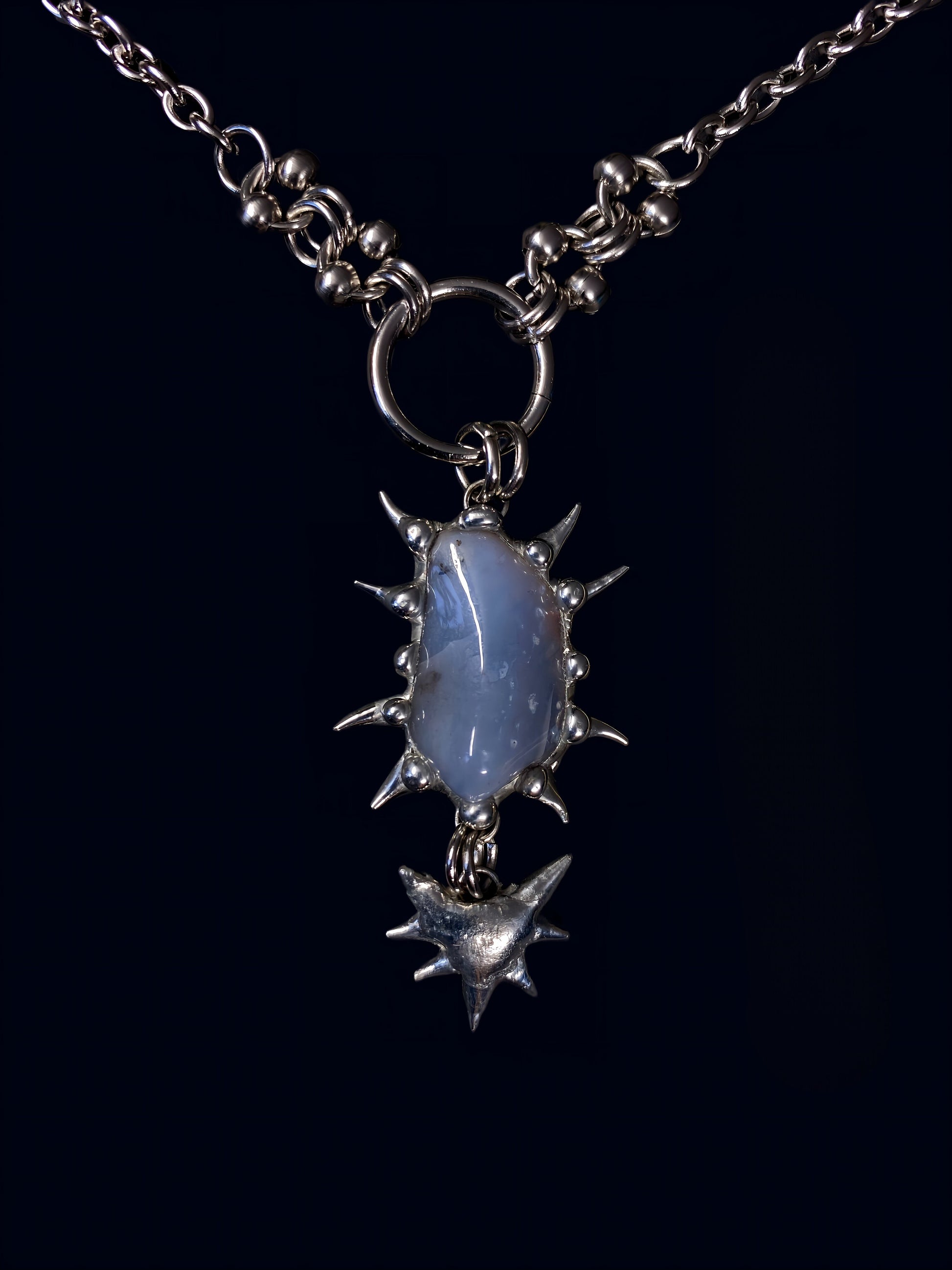 product photo of blue chalcedony gemstone held inside a hand-formed spiked frame made with soft solder. hanging from big o-ring attached to stainless steel link chain. 