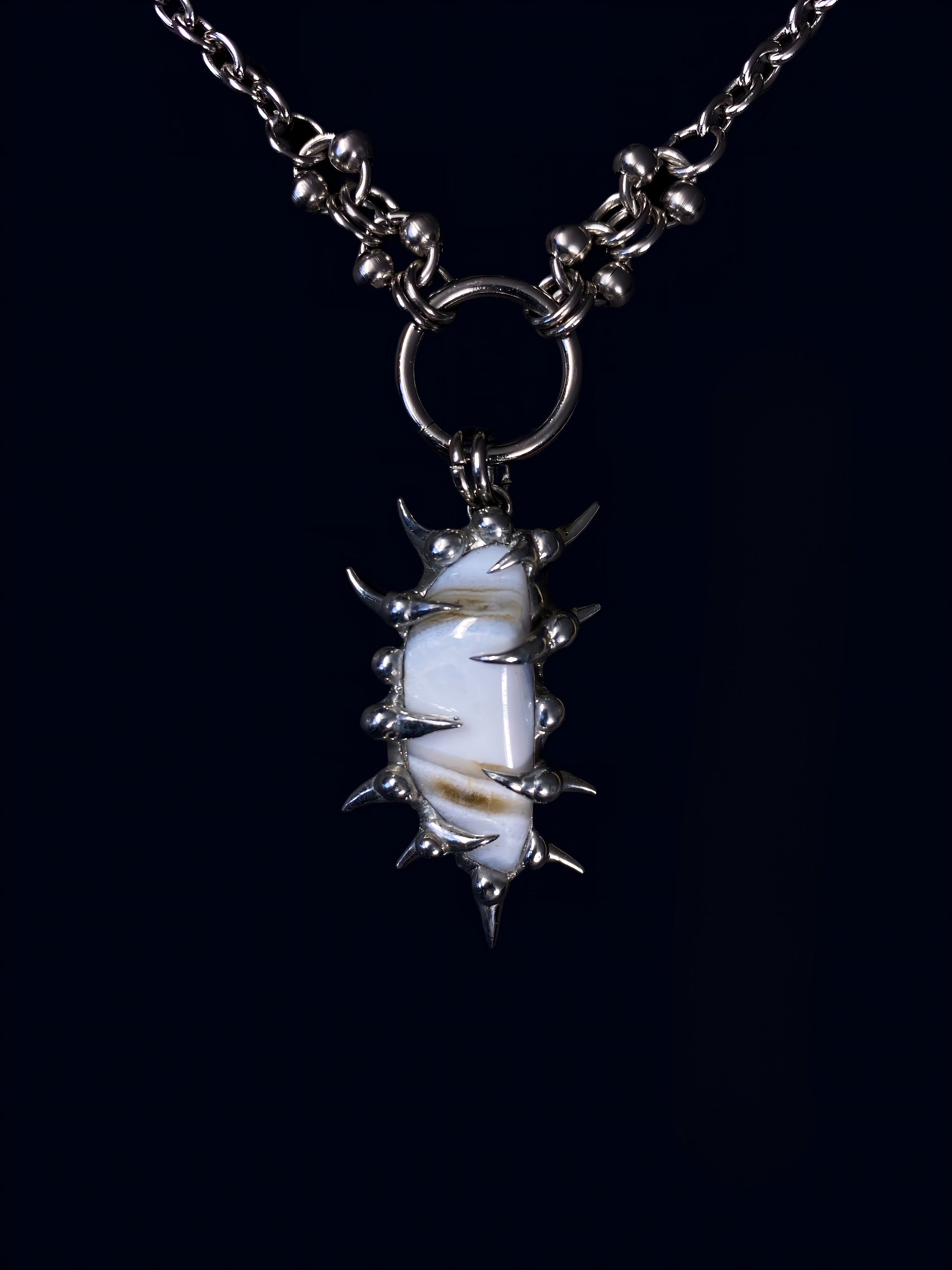 product photo of white calcite gemstone held inside a hand-formed spiked frame made with soft solder. soft brown stripes on the stone. hanging from big o-ring attached to stainless steel link chain. 