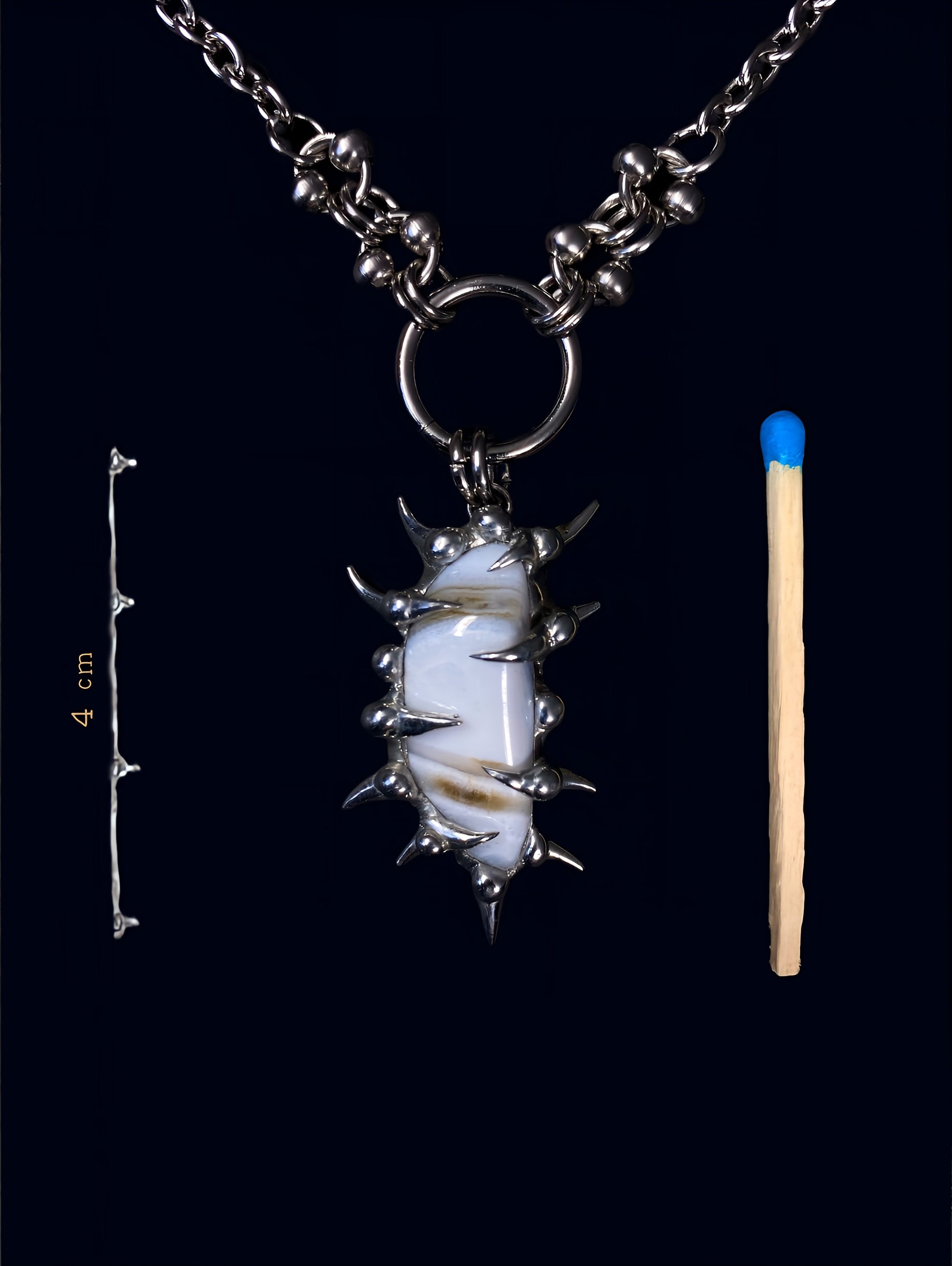 product photo of white calcite gemstone held inside a hand-formed spiked frame made with soft solder. soft brown stripes on the stone. hanging from big o-ring attached to stainless steel link chain. next to it a match to compare size and a measurement of 4 cm. 