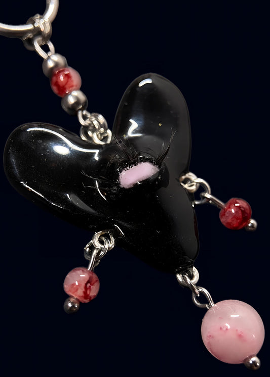 close up product photo of the center pendant from handmade black heart swarm necklace. black heart pendant with one pink eye and small lashes, thrifted red and pink pearls dangling from the heart. made from polymer clay.