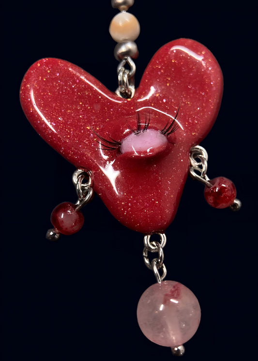close up product photo of the center pendant from handmade glittery heart swarm necklace. red heart pendant with one pink eye and small lashes, thrifted red, pink and white pearls dangling from the heart. made from polymer clay.