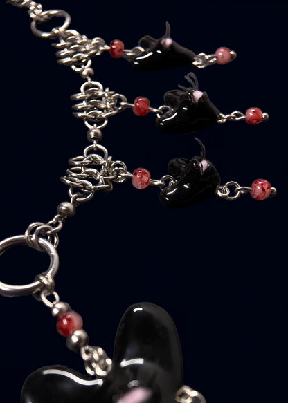 close up product photo of chainmail and pearl part from handmade black heart swarm necklace. small black heart pendant with one pink eye and small lashes, thrifted red pearls dangling from the heart and chainmail necklace. more of the necklace out of focus. 