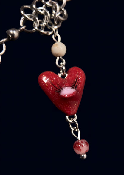 close up product photo of one small pendant from handmade glittery heart swarm necklace. small red heart pendant with one pink eye and small lashes, thrifted red and white pearls dangling from the heart and chainmail necklace. 