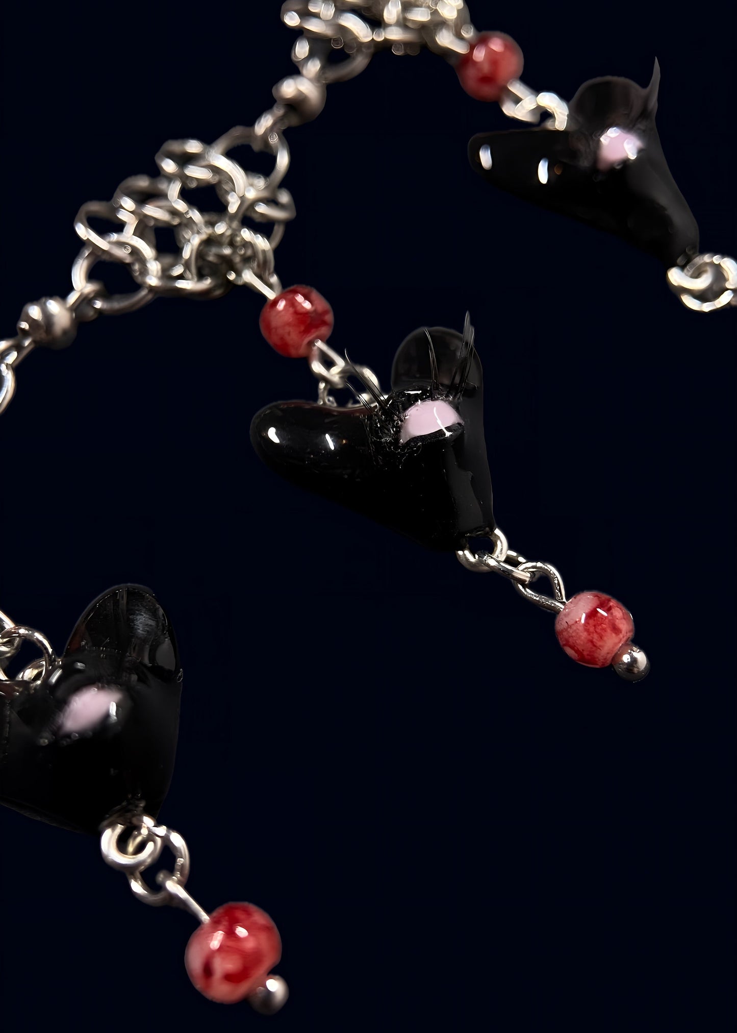 close up product photo of the smaller pendants from handmade black heart swarm necklace. focus on small black heart pendant with one pink eye and small lashes, thrifted red pearls dangling from the heart and chainmail necklace. 2 more small pendants can be seen out of focus.