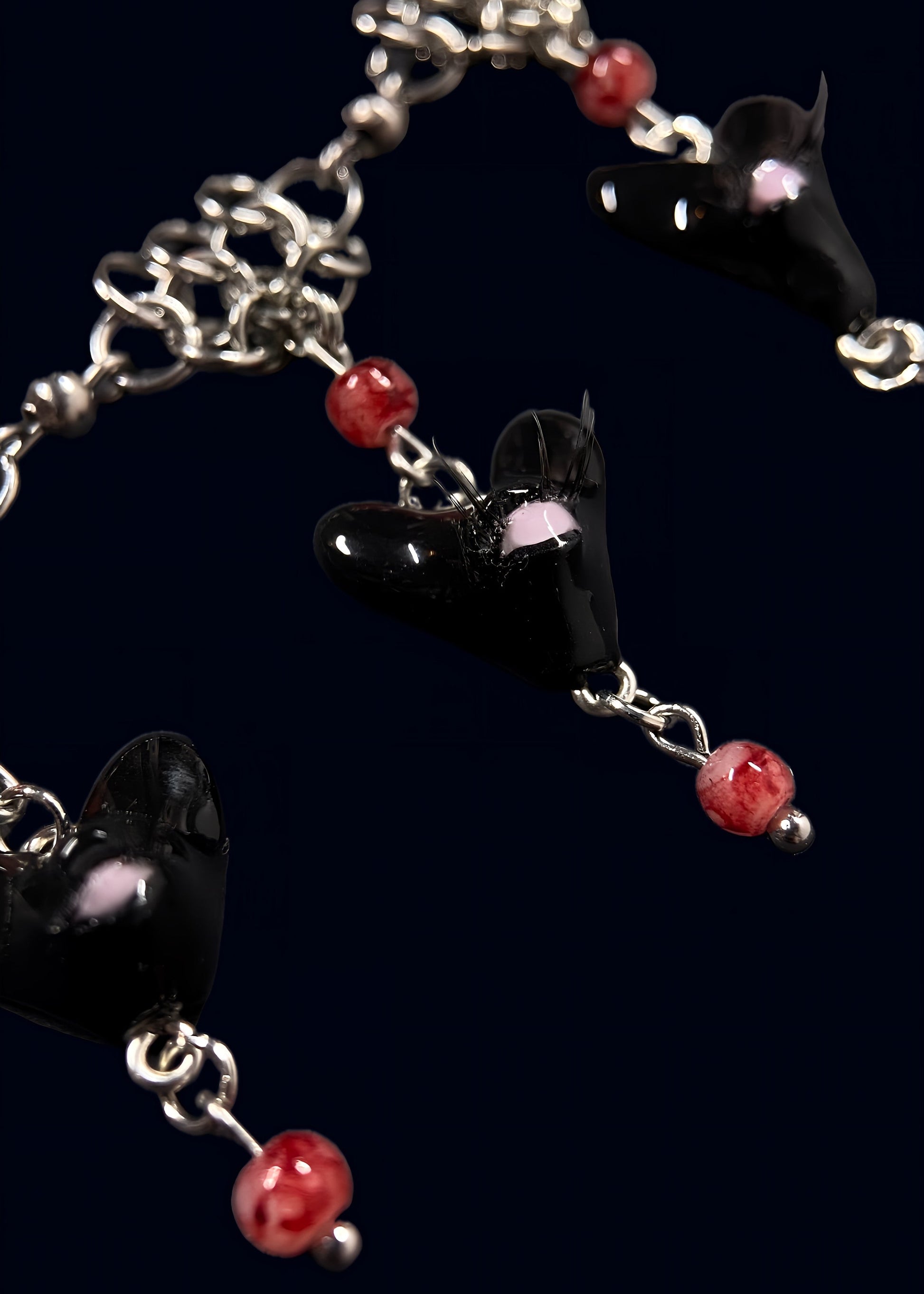 close up product photo of the smaller pendants from handmade black heart swarm necklace. focus on small black heart pendant with one pink eye and small lashes, thrifted red pearls dangling from the heart and chainmail necklace. 2 more small pendants can be seen out of focus.