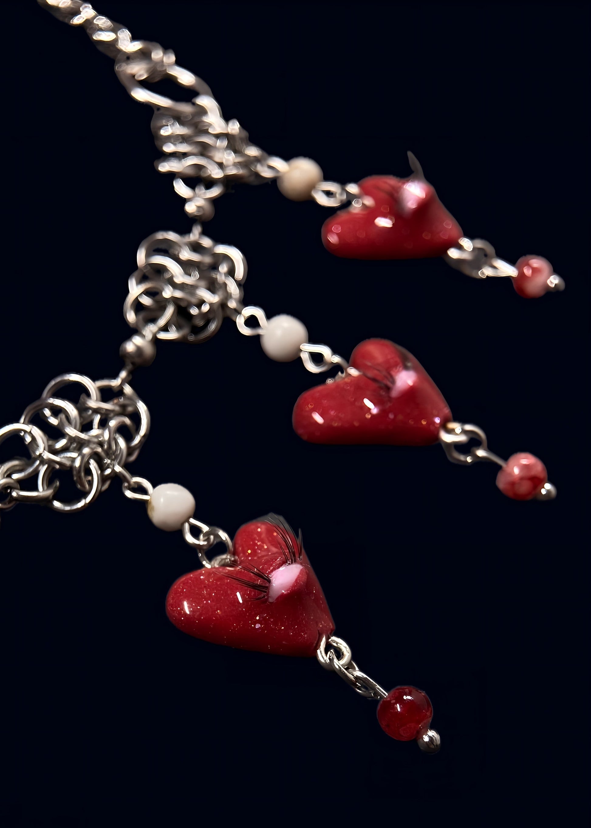 close up product photo of the smaller pendants from handmade glittery heart swarm necklace. focus on small red heart pendant with one pink eye and small lashes, thrifted red and white pearls dangling from the heart and chainmail necklace. 2 more small pendants can be seen out of focus.