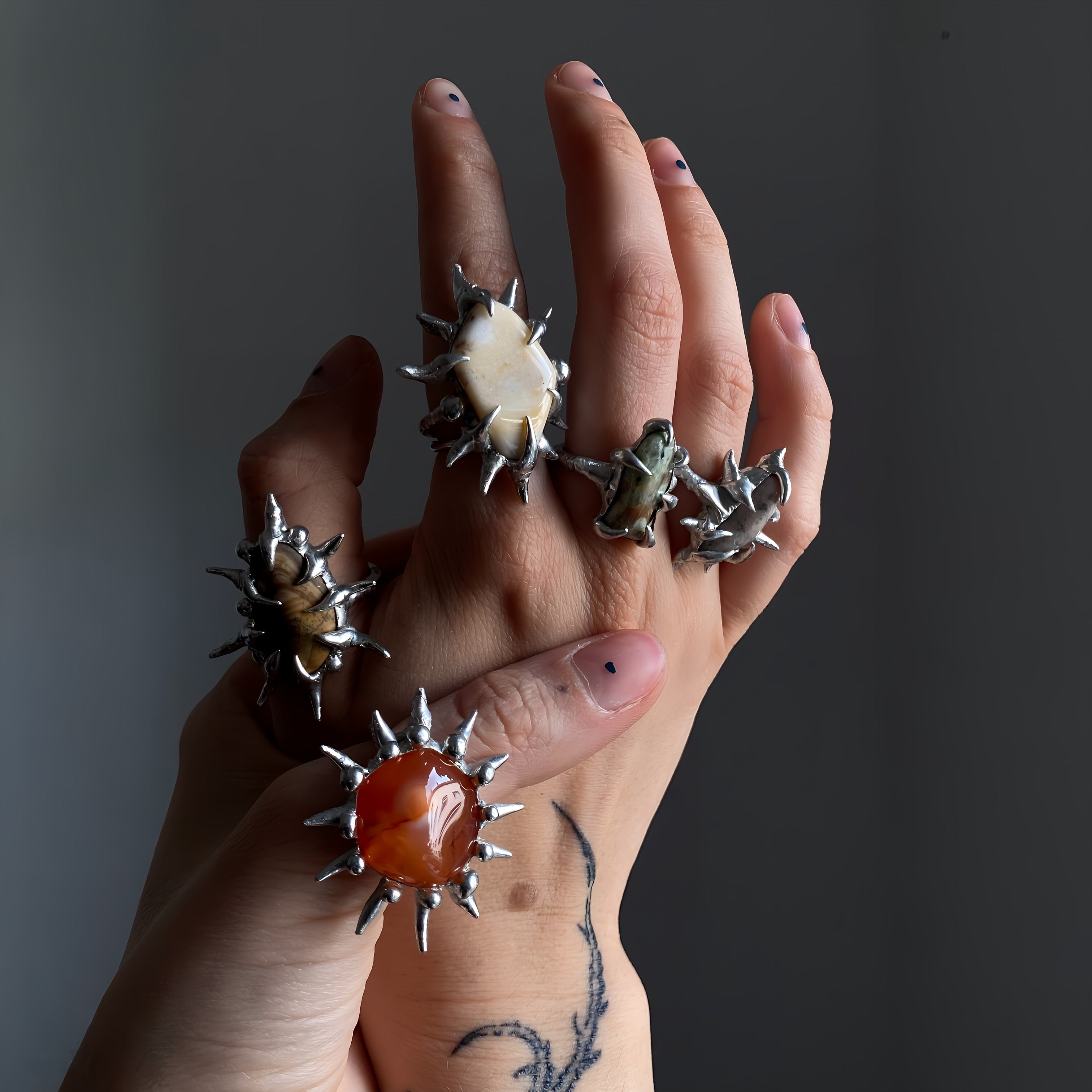 two tattooed hands holding each other while wearing 5 spiky soft soldered rings with different gemstones from latest ring collection.