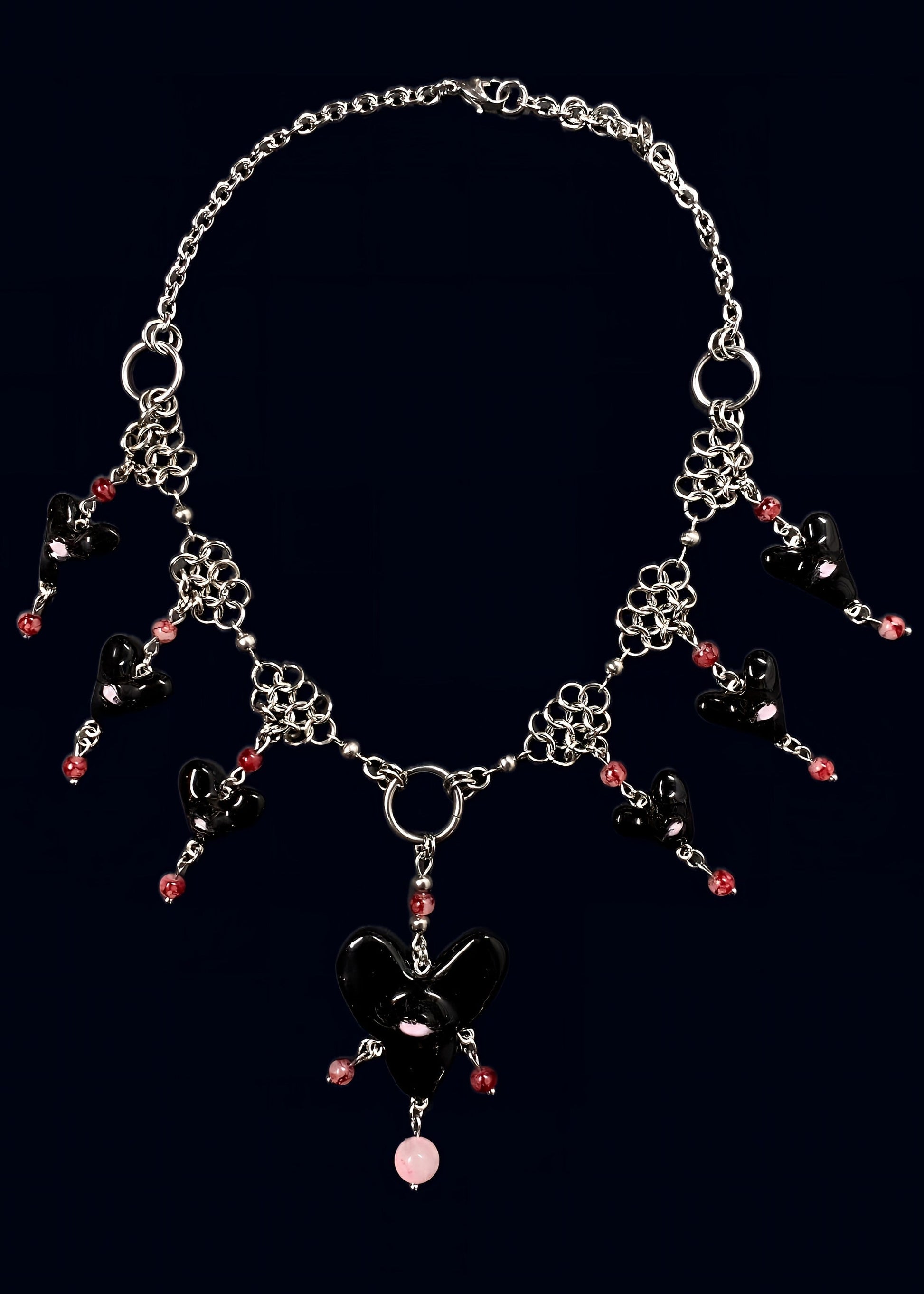 handmade black polymer clay heart swarm necklace: central large red heart pendant with one eye and lashes, six smaller black hearts clustered around it, thrifted red pearls dangling from each heart and woven into stainless steel chainmail necklace, glossy uv resin finish, queer fairycore alt-fashion jewelry by softedgez from berlin