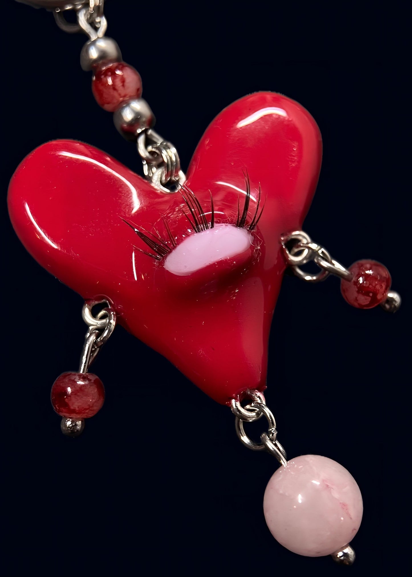 close up product photo of central large red heart pendant with one eye and lashes, thrifted red pearls dangling from each side of the heart and a bigger thrifted pink pearl hanging below. 