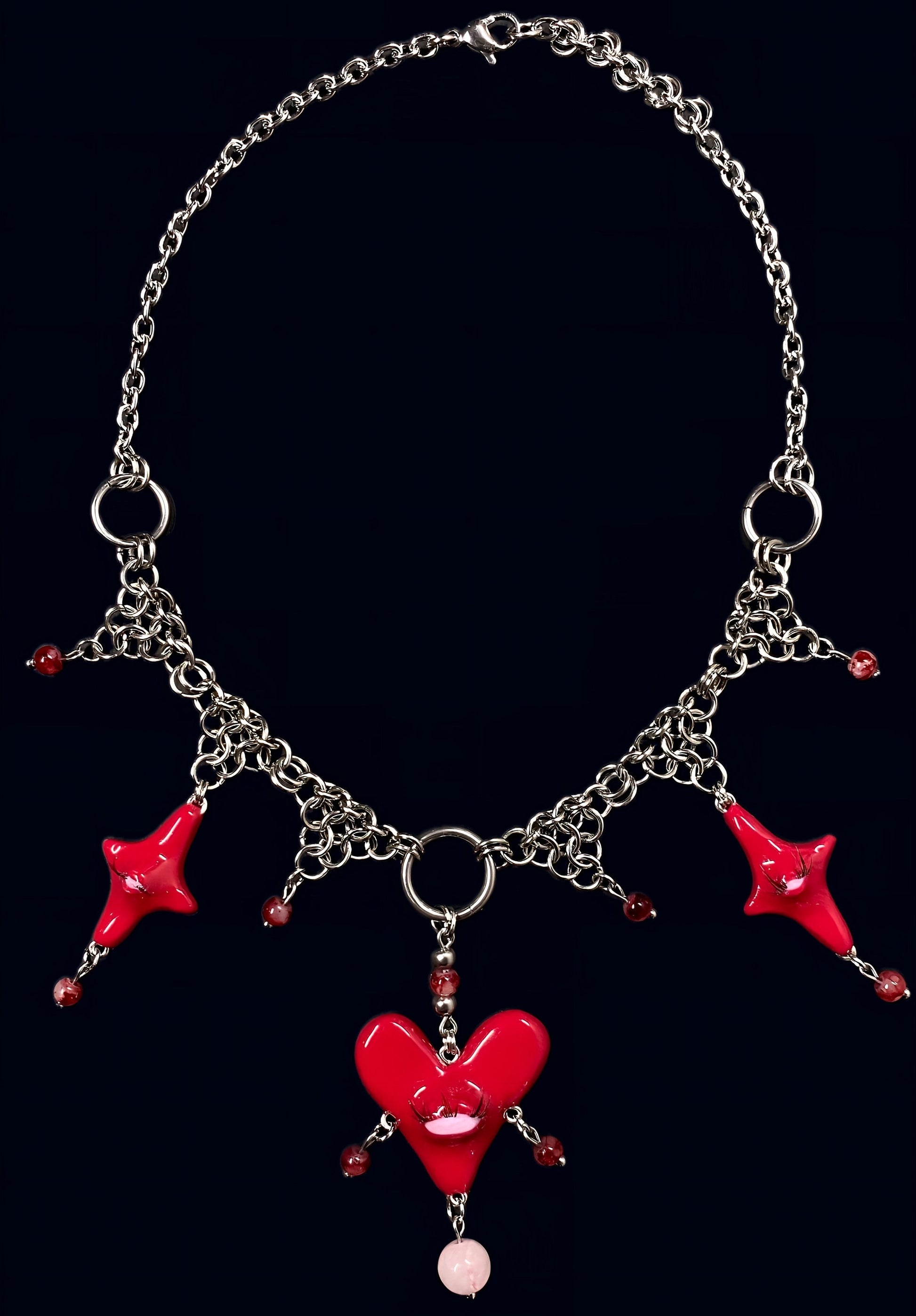 product photo of red heart creature necklace. shows a central large red heart pendant with one eye and lashes, 1 smaller red stars on each side around it. thrifted red pearls dangling from each heart and woven into stainless steel chainmail necklace, glossy uv resin finish, queer fairycore alt-fashion jewelry by softedgez from berlin
