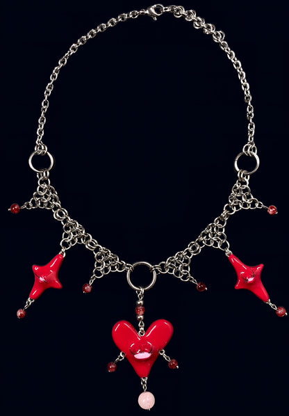 product photo of red heart creature necklace. shows a central large red heart pendant with one eye and lashes, 1 smaller red stars on each side around it. thrifted red pearls dangling from each heart and woven into stainless steel chainmail necklace, glossy uv resin finish, queer fairycore alt-fashion jewelry by softedgez from berlin